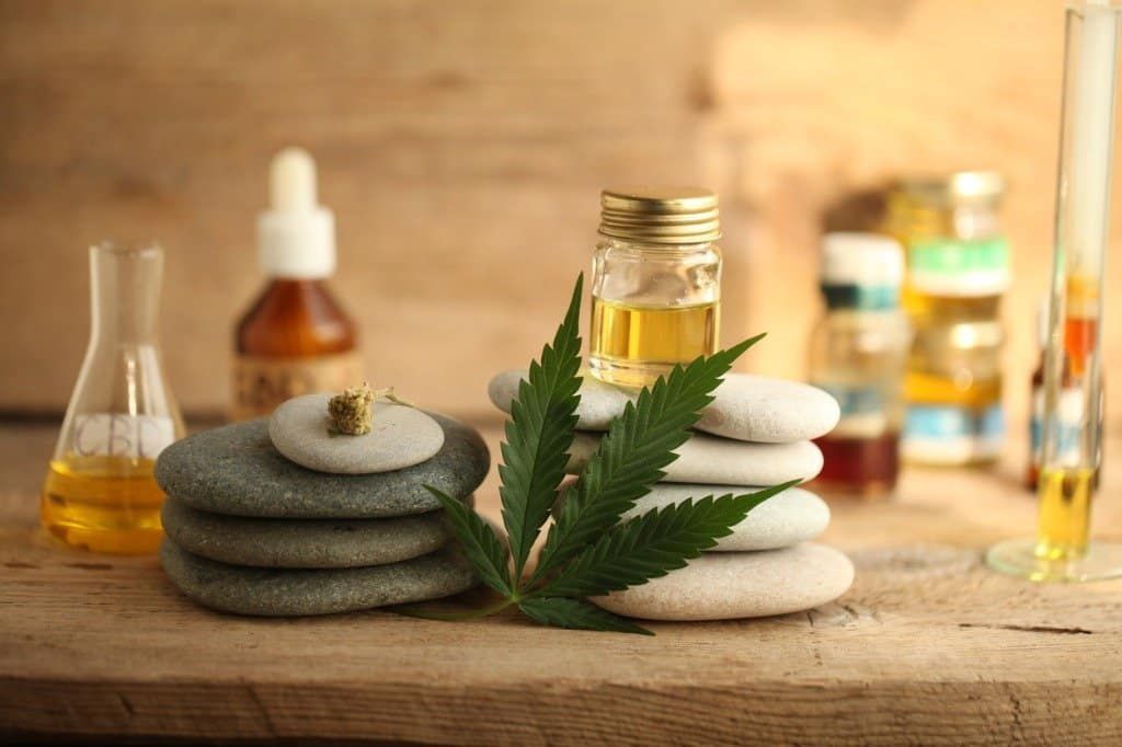 cannabis-leaf-resting-against-pile-of-stones-on-table-with-vials-and-bottles-of-cbd-and-other-essential-oils